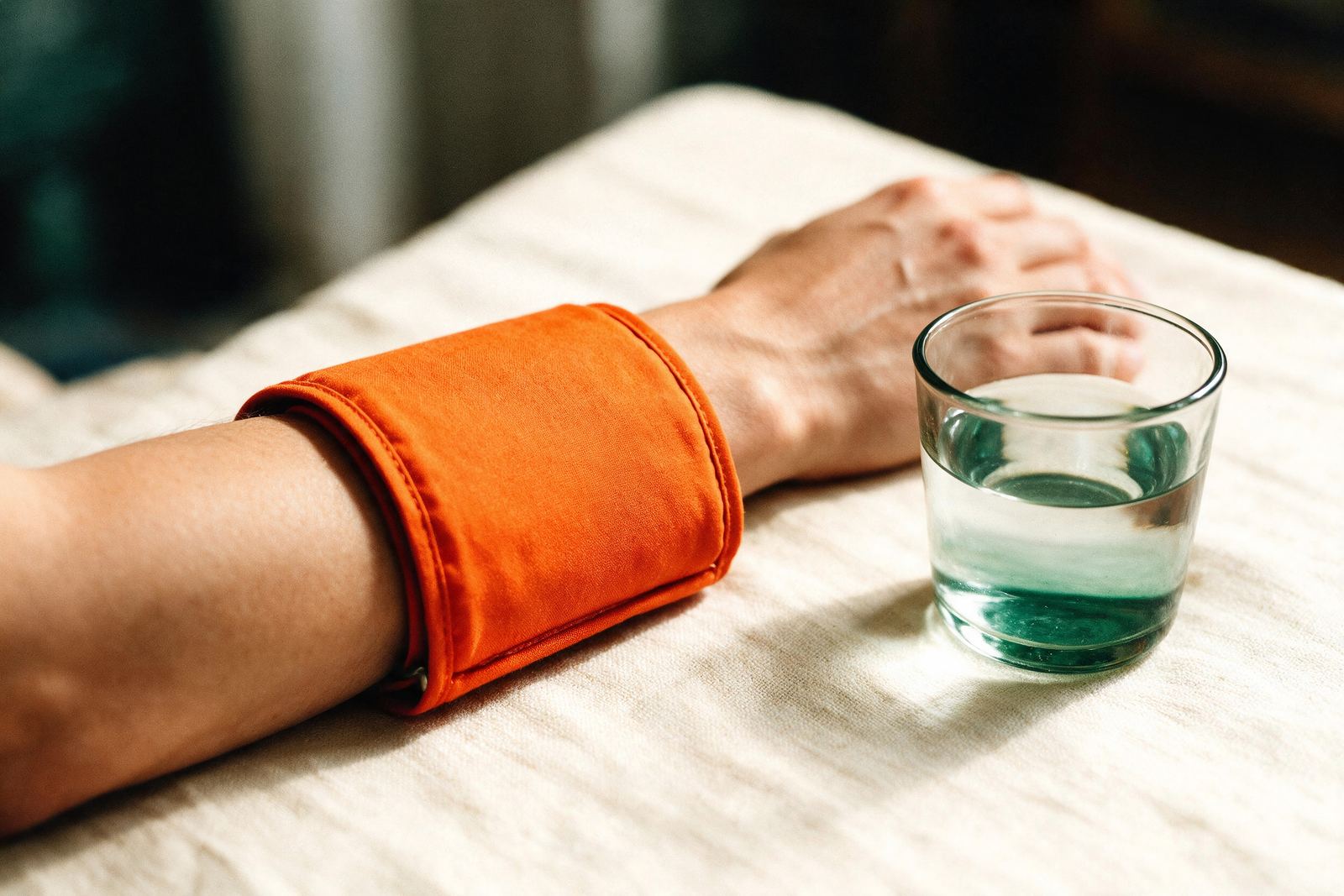 A blood pressure cuff on a forearm resting on cream linen beside a glass of water.