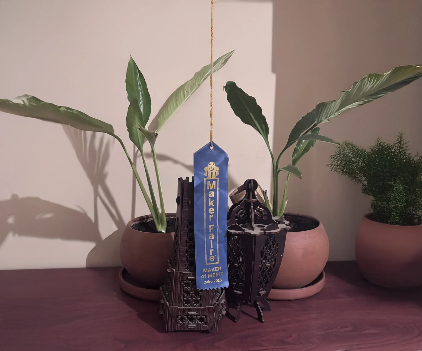 The blue Maker of Merit ribbon from Maker Faire Cairo 2016, hanging beside the laser-cut mashrabiya lantern and tower it was awarded for.